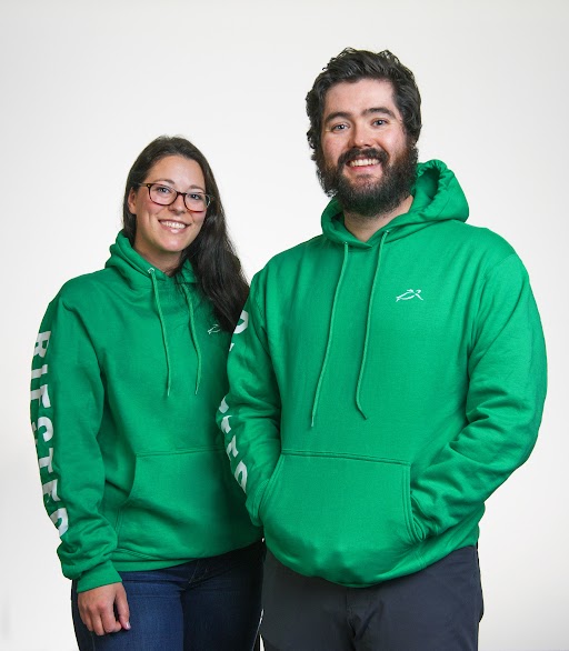 Female and male young professionals stand smiling in green RIESTER branded sweatshirts.
